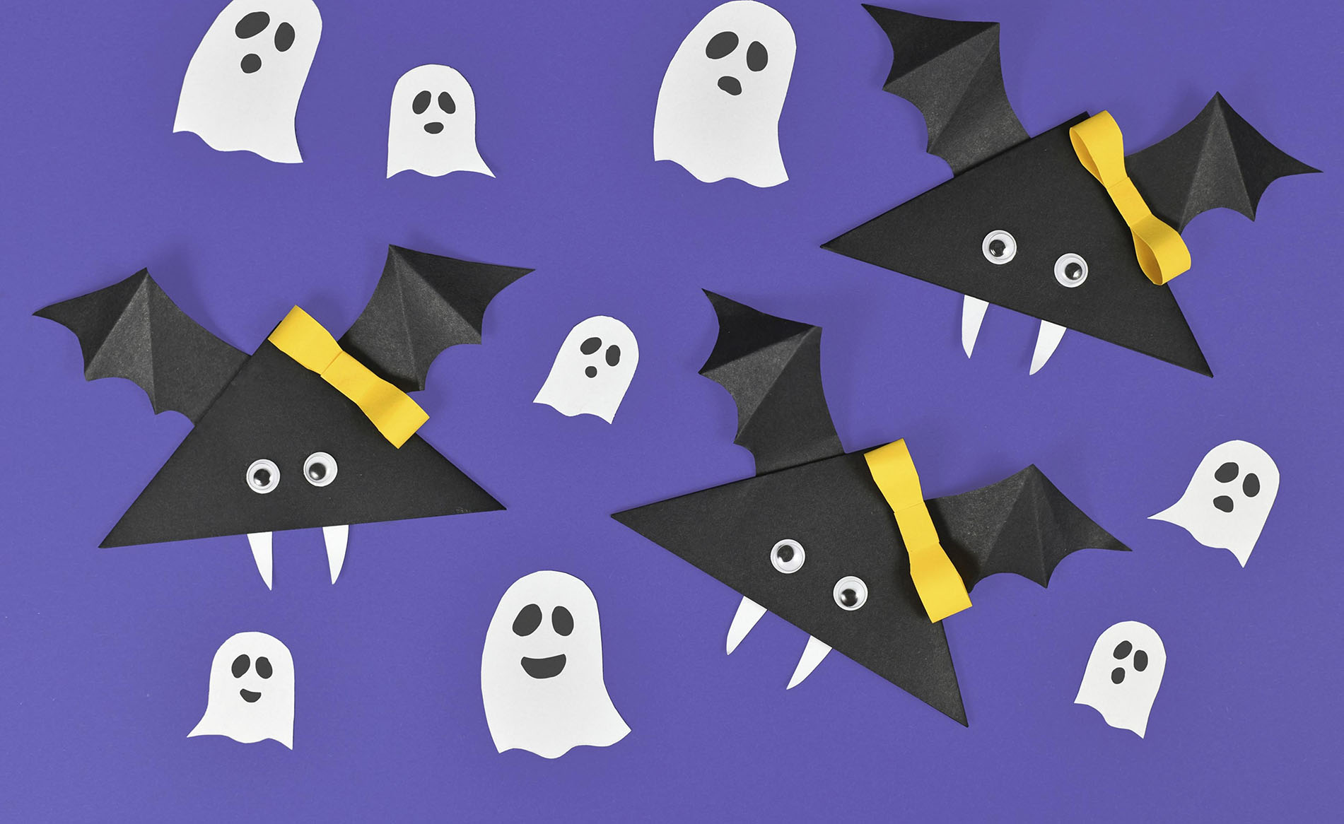 Paper vampire bats with funny googly eyes and ribbons and ghosts on purple Halloween background,