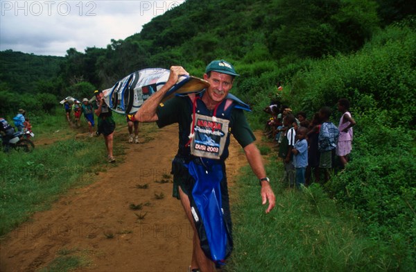 senior man with double canoe on shoulder walks toward camera, lo