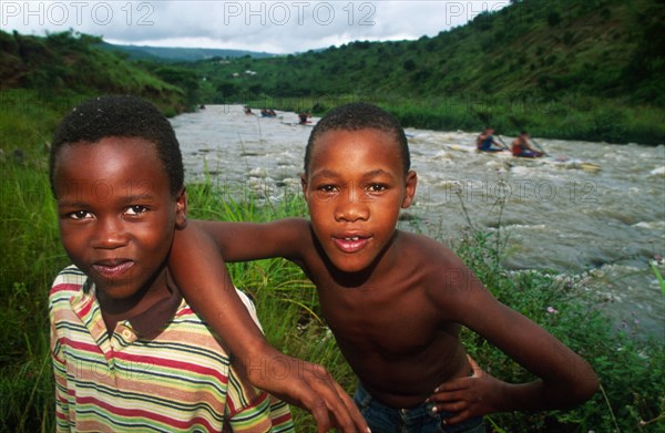 two boys on bank of river look into camera, paddlers behind on r