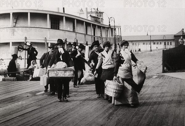 Imigrants to US landing at Ellis Island circa 1900. They head for the processing centre carrying paper with entry number which they hope will soon be traded for a visa.