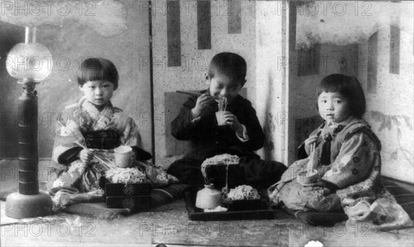 Three Japanese children seated on cushions on the floor,  eating noodles with chopsticks