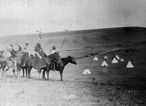 Four Atsina Indians on horseback overlooking tepees in valley beyond