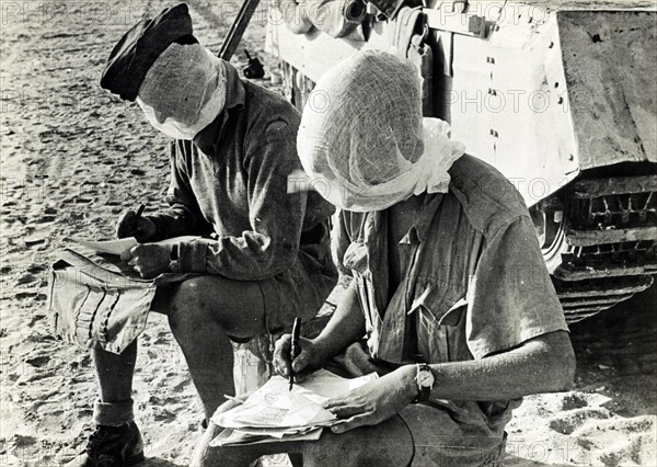 Photographic print of British World War Two soldiers with their heads covered, writing letters beside a tank