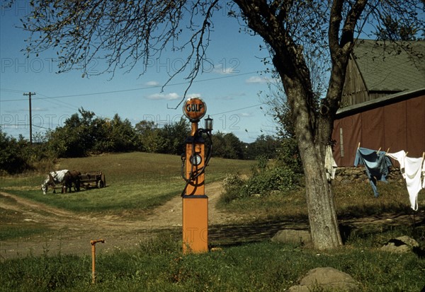 Gas pump with clothesline, barn and horse-drawn wagon in background.