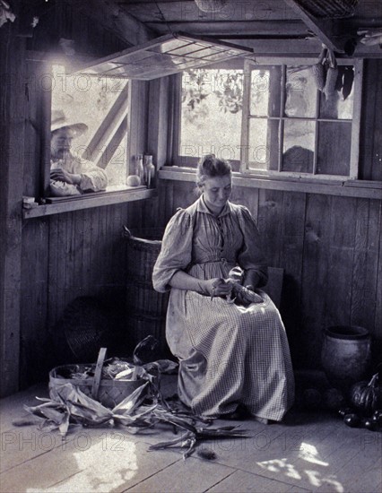 Photograph shows rustic scene of a woman seated while a man leans on the window sill.