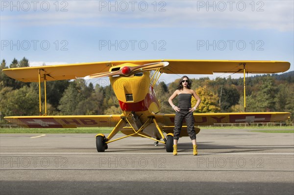 Young woman with sunglasses in overall and boots posing in front of double-decker airplane