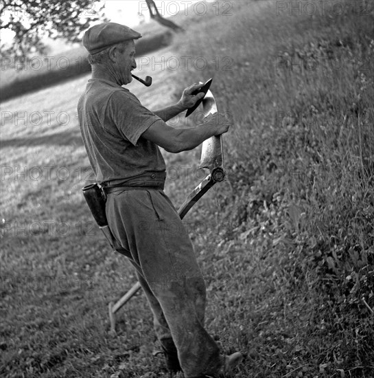 Man with a pipe sharpened in his mouth sharpens a scythe with the help of a grindstone