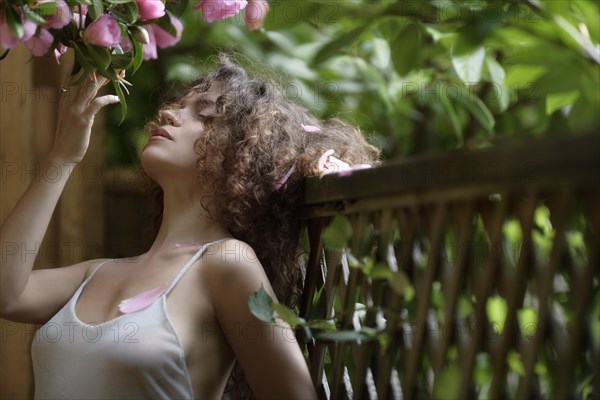 Sensual portrait of a young beautiful woman outdoors leaning against a fence touching blooming pink camellia tree flowers