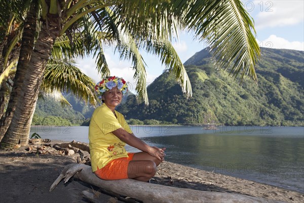 Old woman with wreath of flowers sitting under palm trees on tree trunk