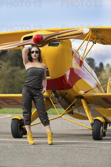 Young woman with sunglasses in overall and boots posing in front of double-decker airplane