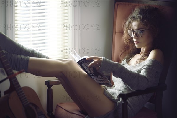 Young woman lying lascivious in a chair and reading a book by a window in daylight