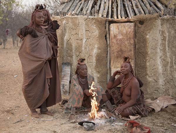 Himba women sit early in the morning in front of the mud hut at the burning fire