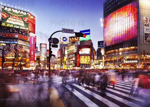 Crowd of people crossing Meiji dori street at Shibuya station busy intersection lit with colorful signs and ads in the evening rush hour