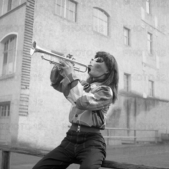 Man with shirt and mask sits on a wooden railing and plays trumpet