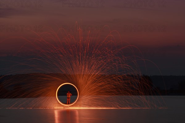 A man stands on a platform in a lake and creates a hoop of fire with glowing steel wool around him