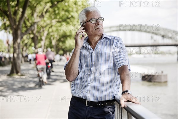 Grauhaariger Senior calls with his smartphone on Cologne's Rhine bank promenade in front of Hohenzollern Bridge