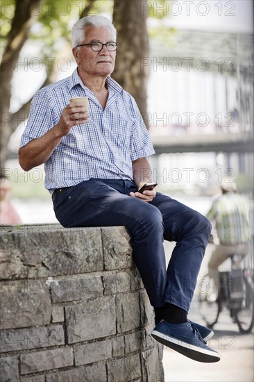 Senior sits with his Smartphone and a Coffe to go on a wall at Cologne's Rhine bank Promenade