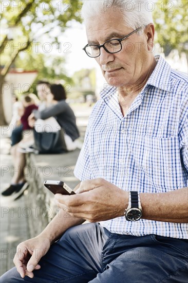 Grey-haired senior sits on a wall with his smartphone in his hand