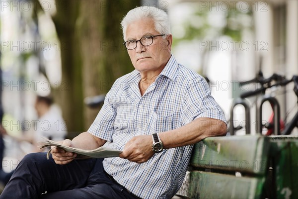 Grauhaariger senior sits in Cologne's Old Town on a bank and reads a newspaper