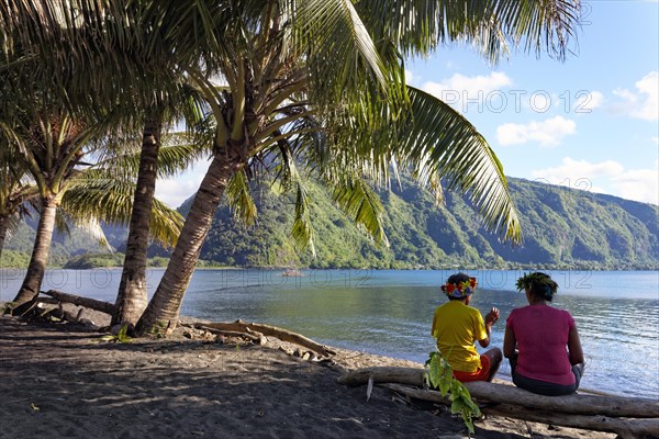 Two women with wreath of flowers sitting under palm trees on tree trunk