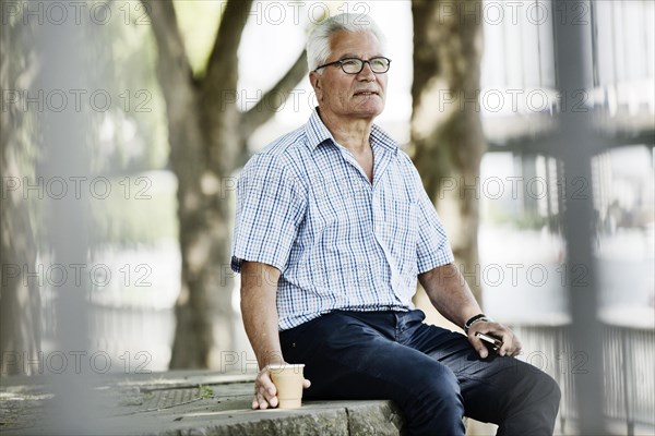 Senior sits with his Smartphone and a Coffe to go on a wall at Cologne's Rhine bank Promenade