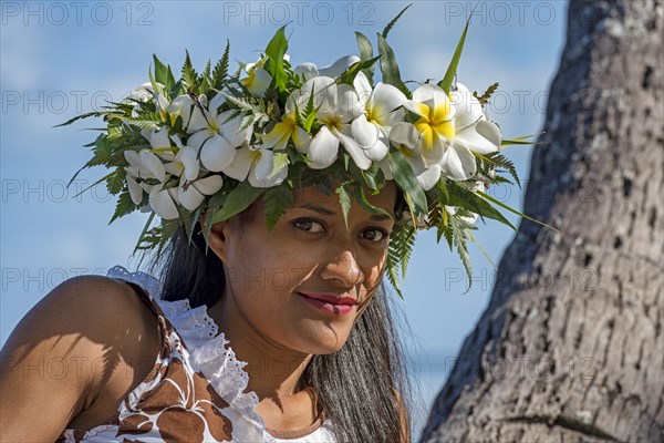 Young woman with flower wreath from Frangipani leaning on palm trunk