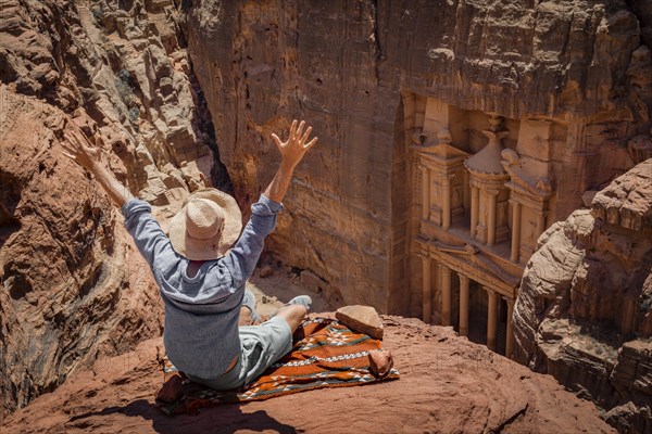 Tourist with sun hat sits with raised arms on carpet and looks from above into the canyon Siq