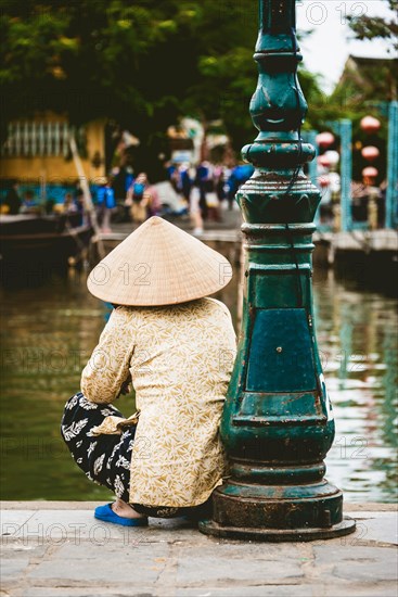 Native woman with straw hat sits on the shore of the Thu Bon River