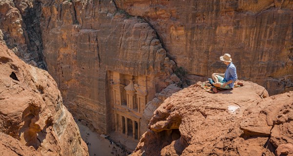 Tourist with sun hat sits on rocks and looks from above into the canyon Siq