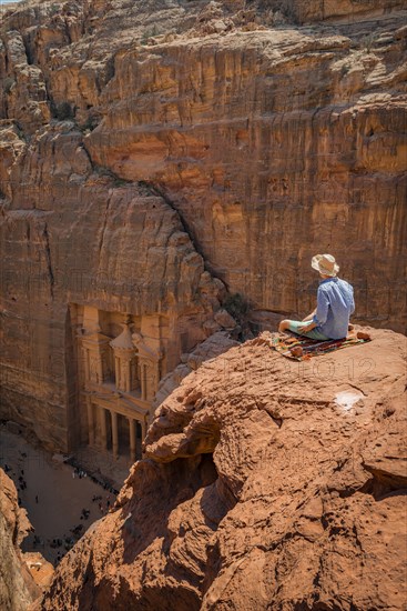 Tourist with sun hat sits on rocks and looks from above into the canyon Siq