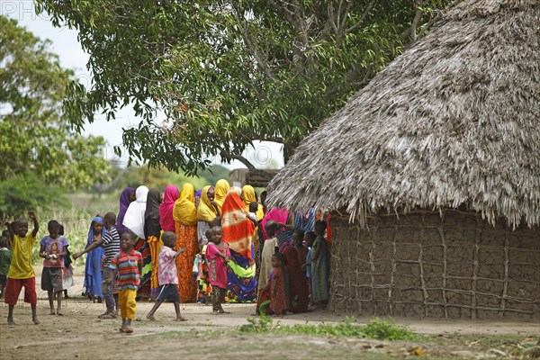 Women and children in colourful clothing next to mud hut