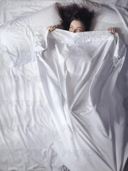 Cute young woman lying in bed covered with a bedsheet up to her eyes peeking from under a blanket