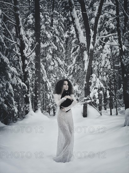 Beautiful woman in white long dress walking in the snow with a black cat in her hands