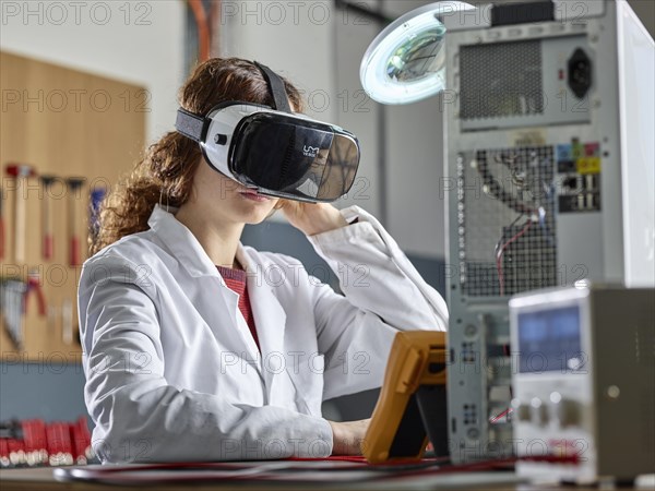 Woman with VR goggles and lab coat sitting in front of PC open in an electronics laboratory