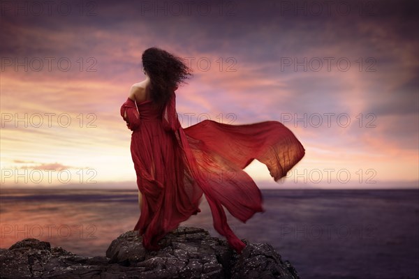Young woman in red long summer dress flying in the wind standing on a rocky shore