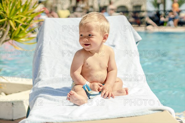 Blond toddler sitting on the sun bed by swimming pool in the resort