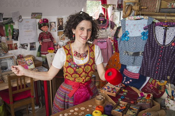 Button maker in colorful dirndl with Collier button trimmings in handicraft studio working with red yarn and utensils for Posamentenknopfe