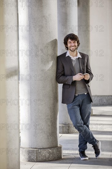 Young man laughingly leans against column with smartphone in hand