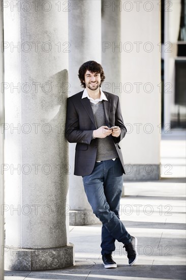 Young man laughingly leans against column with smartphone in hand