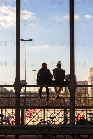 Two young women sitting on the balustrade of the Hackerbrucke bridge over the railway tracks looking into the distance