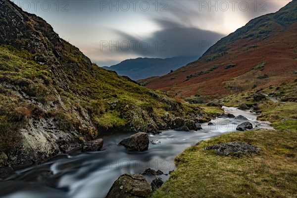 Brook run in autumnal hilly landscape with cloudy sky