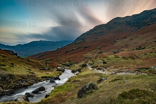 Brook run in autumnal hilly landscape with cloudy sky