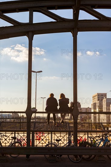 Two young women sitting on the balustrade of the Hackerbrucke bridge over the railway tracks looking into the distance
