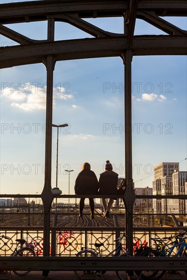 Two young women sitting on the balustrade of the Hackerbrucke bridge over the railway tracks looking into the distance