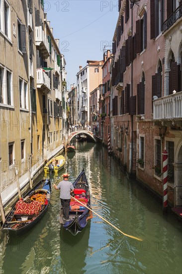 Gondolier steering gondola with tourists on narrow canal with historical buildings