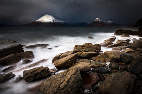 Big rocks in the water of the North Sea with snow-covered Cullin mountains in the background