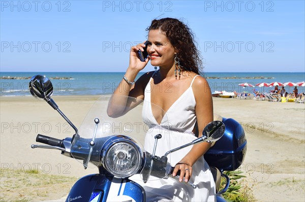 Woman in white dress and decorative earrings with cell phone on Vespa Primavera scooter on the beach