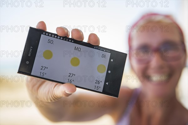 Laughing woman on the beach happy about sunny weather in the weather forecast on her smartphone