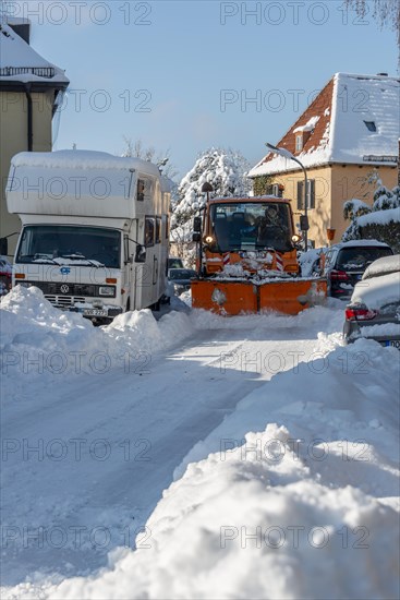Snowplough drives through narrow road with snow-covered parked cars