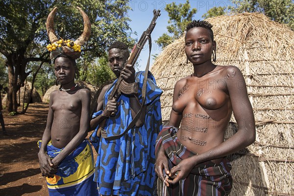 Young girls with decorative scars and man with Kalashnikov in front of hut